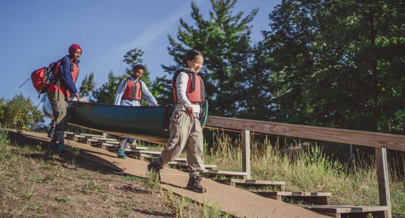 Three people wearing life jackets carry a canoe down a concrete slope, presumably toward water. 